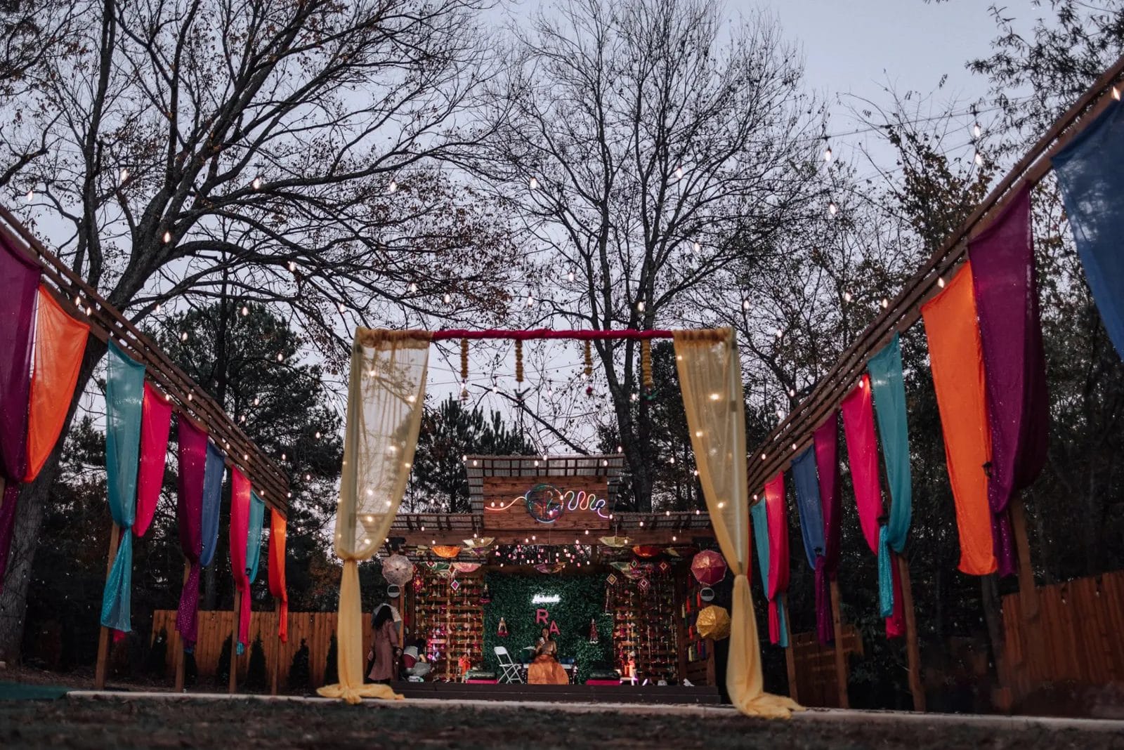 Outdoor wedding altar with colorful drapes in orange, pink, and blue hanging on wooden structures. The altar is adorned with greenery and a backdrop of trees. String lights are visible above, and it is set in a wooded area at twilight. Huntersville event venue
