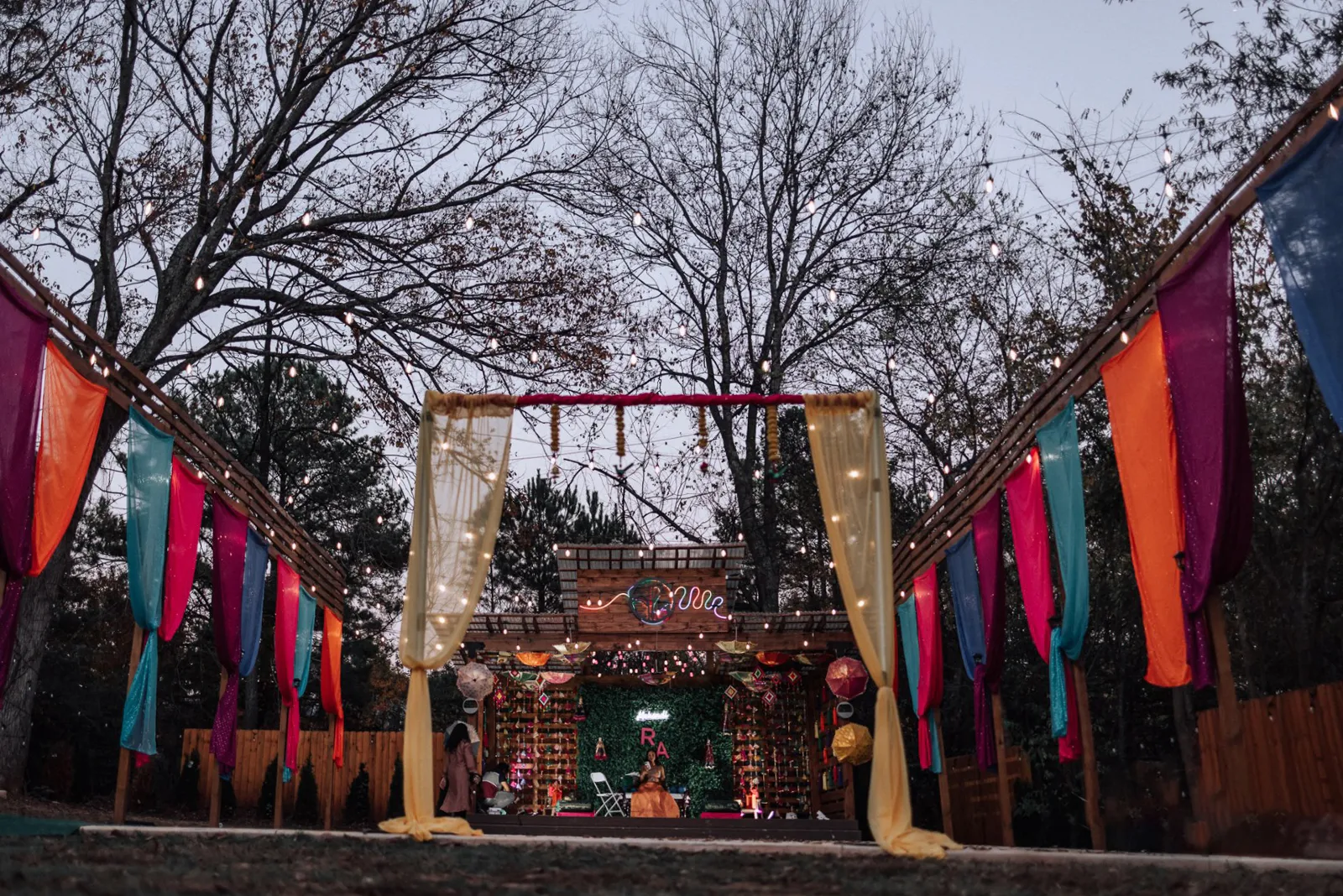 A Beautiful New Addition to Our Outdoor Event Venue with Stage in Huntersville, NC 2 Outdoor wedding altar with colorful drapes in orange, pink, and blue hanging on wooden structures. The altar is adorned with greenery and a backdrop of trees. String lights are visible above, and it is set in a wooded area at twilight. Huntersville event venue