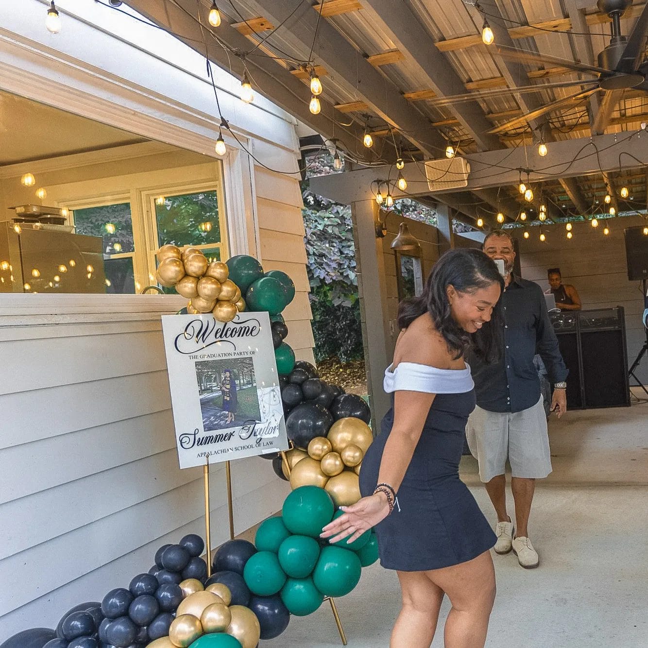 A woman in a black and white off-shoulder dress dances joyfully under a canopy decorated with strings of lights. Nearby is a sign with balloons in black, green, and gold, welcoming guests to a summer party. A DJ is in the background. Huntersville event venue