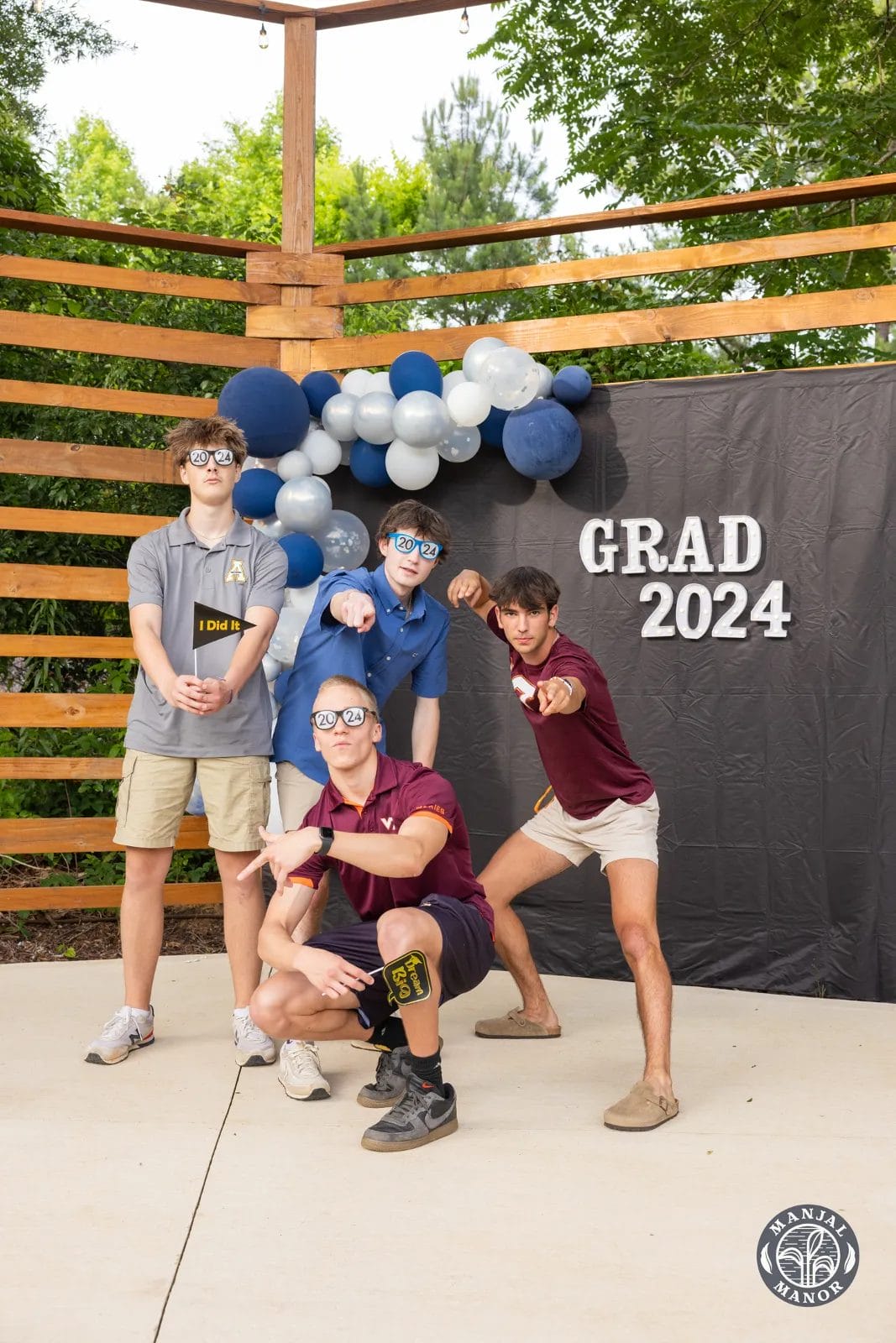 Four young men pose playfully at a "Grad 2024" celebration. They wear casual shirts and shorts, with sunglasses and sneakers. Blue and white balloons decorate the background, with a wooden slat wall and trees visible behind them. Huntersville event venue