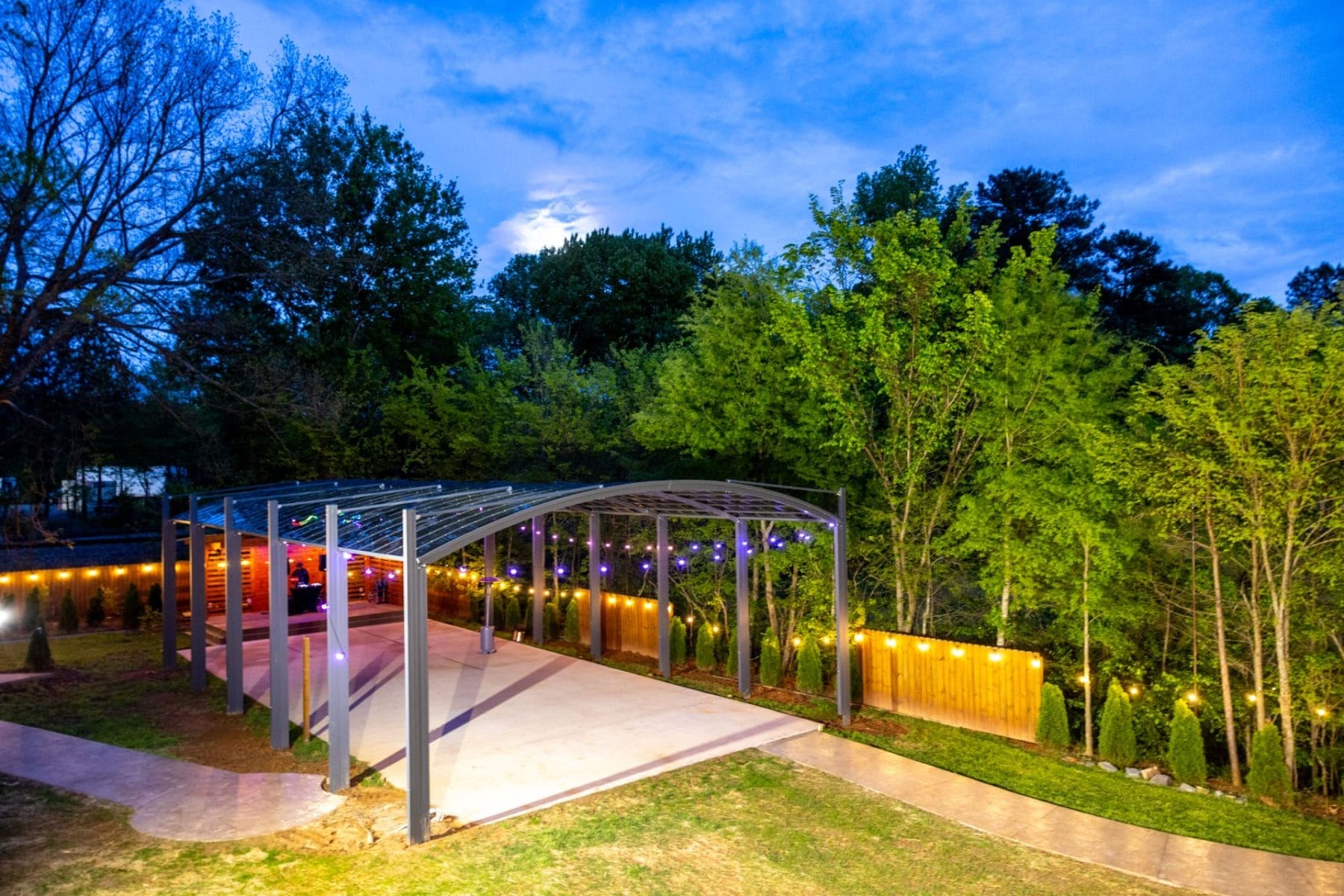 A Beautiful New Addition to Our Outdoor Event Venue with Stage in Huntersville, NC 1 A backyard scene at dusk features a modern pergola with string lights. The area is surrounded by trees and a wooden fence adorned with lights. The sky shows hints of blue, adding to the tranquil ambiance. Huntersville event venue