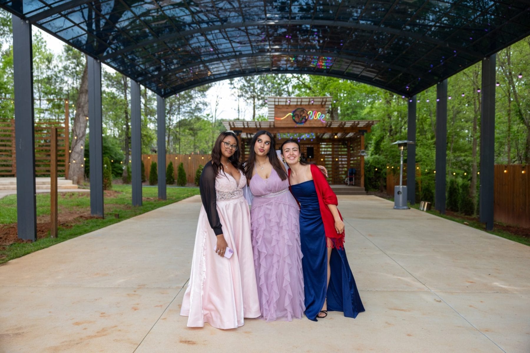 Three women in formal evening gowns stand arm-in-arm under a glass-roofed pavilion. The background features green trees and a wooden structure with the word "belles" in colorful letters, creating a festive atmosphere. Huntersville event venue