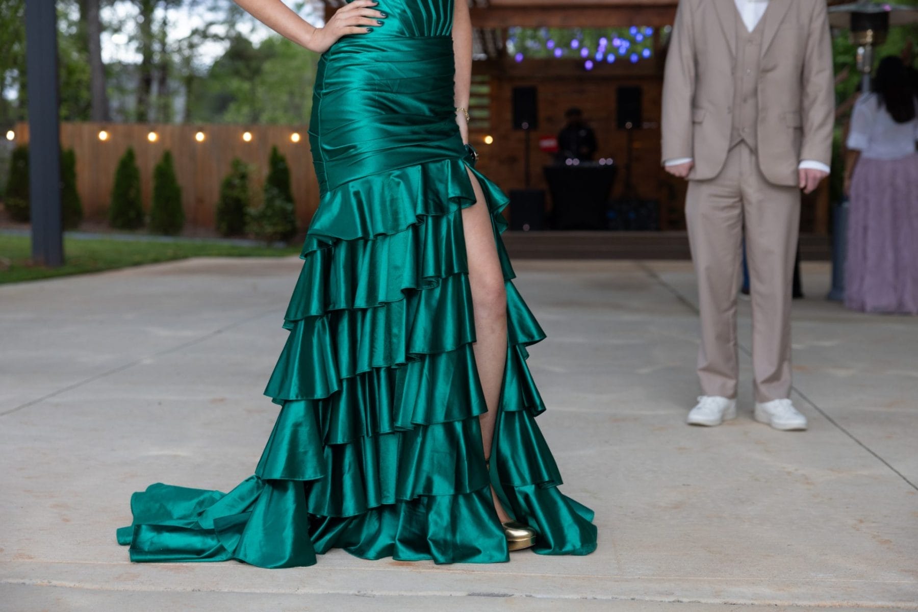 A Beautiful New Addition to Our Outdoor Event Venue with Stage in Huntersville, NC 4 A person in a green, ruffled gown with a thigh-high slit stands outdoors on a concrete floor. Next to them, another person in a light brown suit with white shoes is partially visible. There are twinkling lights and trees in the background. Huntersville event venue