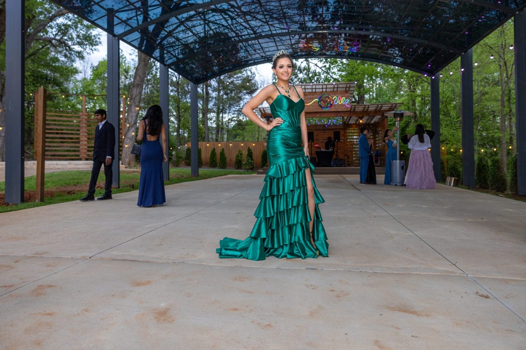 A woman in a vibrant green strapless gown poses confidently under a glass canopy decorated with lights. Several people in formal attire are in the background, and a wooden booth is visible amid lush greenery. Huntersville event venue