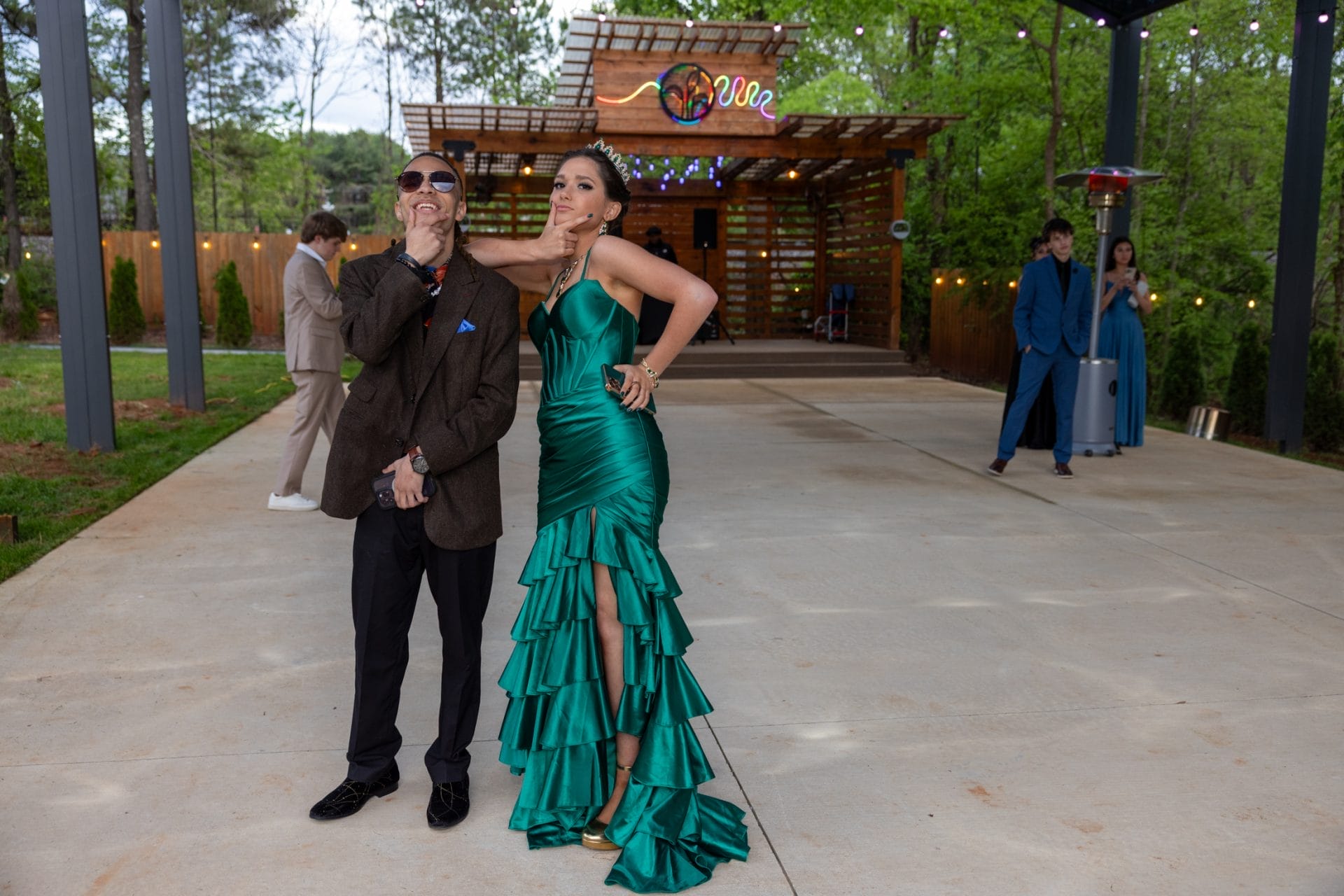 A man and woman strike playful poses in formal attire. The woman wears a green ruffled gown and tiara, and the man wears a suit and sunglasses. A colorful venue with string lights and guests in the background completes the scene. Huntersville event venue