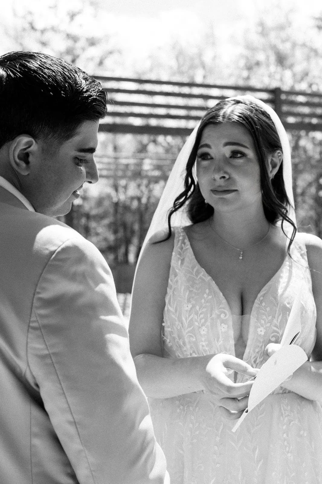 A black and white photo of a couple at their wedding. The bride, in a white dress with a veil, holds a piece of paper and gazes at the groom, who is wearing a light-colored suit. They are outdoors with blurred trees in the background. Huntersville event venue