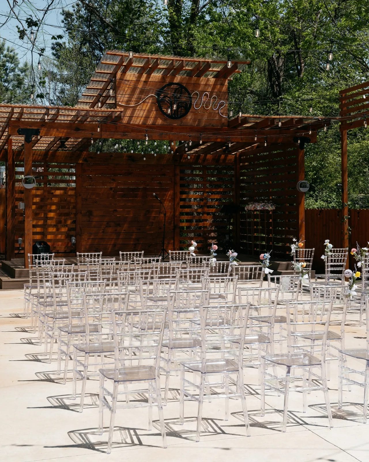 Outdoor wedding venue with rows of translucent chairs facing an empty wooden stage adorned with a circular design. String lights hang above, and trees are visible in the background under a clear blue sky. Huntersville event venue
