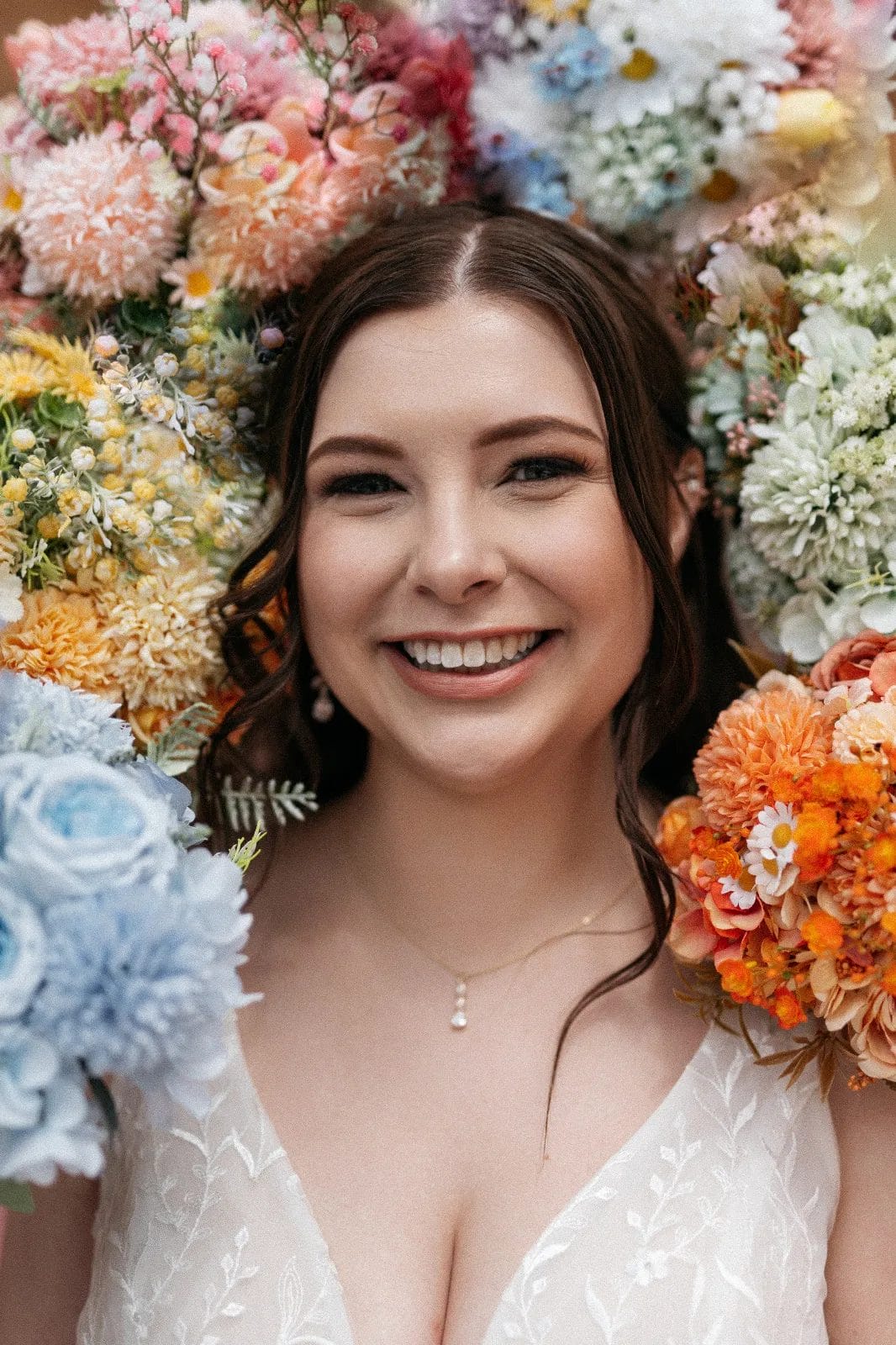 Smiling woman with dark hair stands surrounded by a vibrant assortment of flowers, including orange, blue, and white blooms. She wears a white dress and a delicate necklace. Huntersville event venue