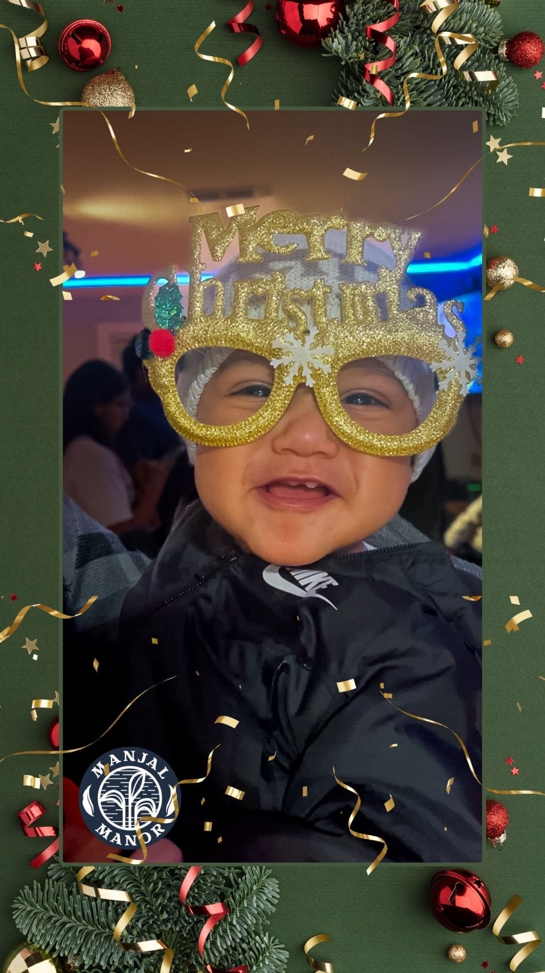 A baby wearing oversized "Merry Christmas" glasses smiles joyfully. The background shows a festive atmosphere with people and decorations. Framing the photo are Christmas-themed borders with ornaments and confetti. Huntersville event venue