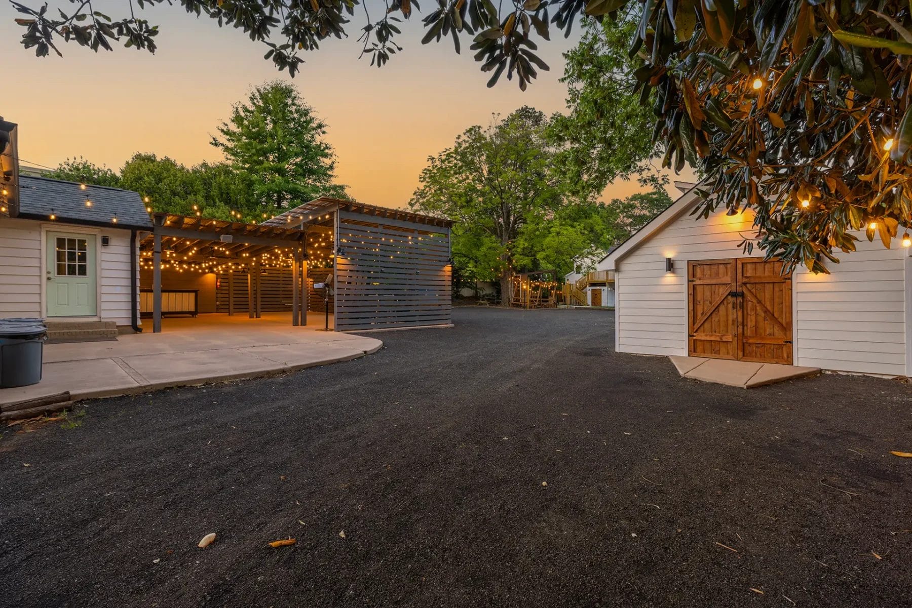 Outdoor area at sunset featuring a small white building with wooden double doors, a covered patio with string lights, a gravel driveway, and trees in the background, creating a warm and inviting atmosphere. Huntersville event venue