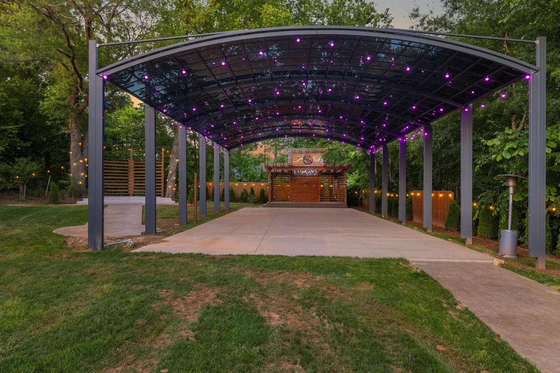 A modern outdoor pavilion with a curved transparent roof and purple string lights, covering a concrete patio, surrounded by green grass, trees, and a wooden structure in the background. Huntersville event venue