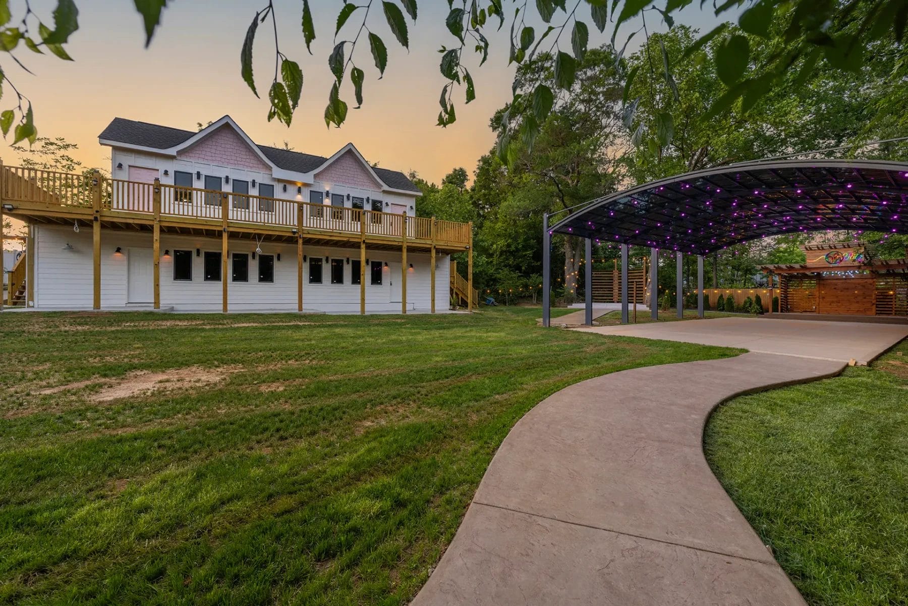 A white two-story house with a large wooden deck overlooks a neatly mowed lawn. A winding concrete path leads to a covered outdoor area with string lights, surrounded by trees at sunset. Huntersville event venue