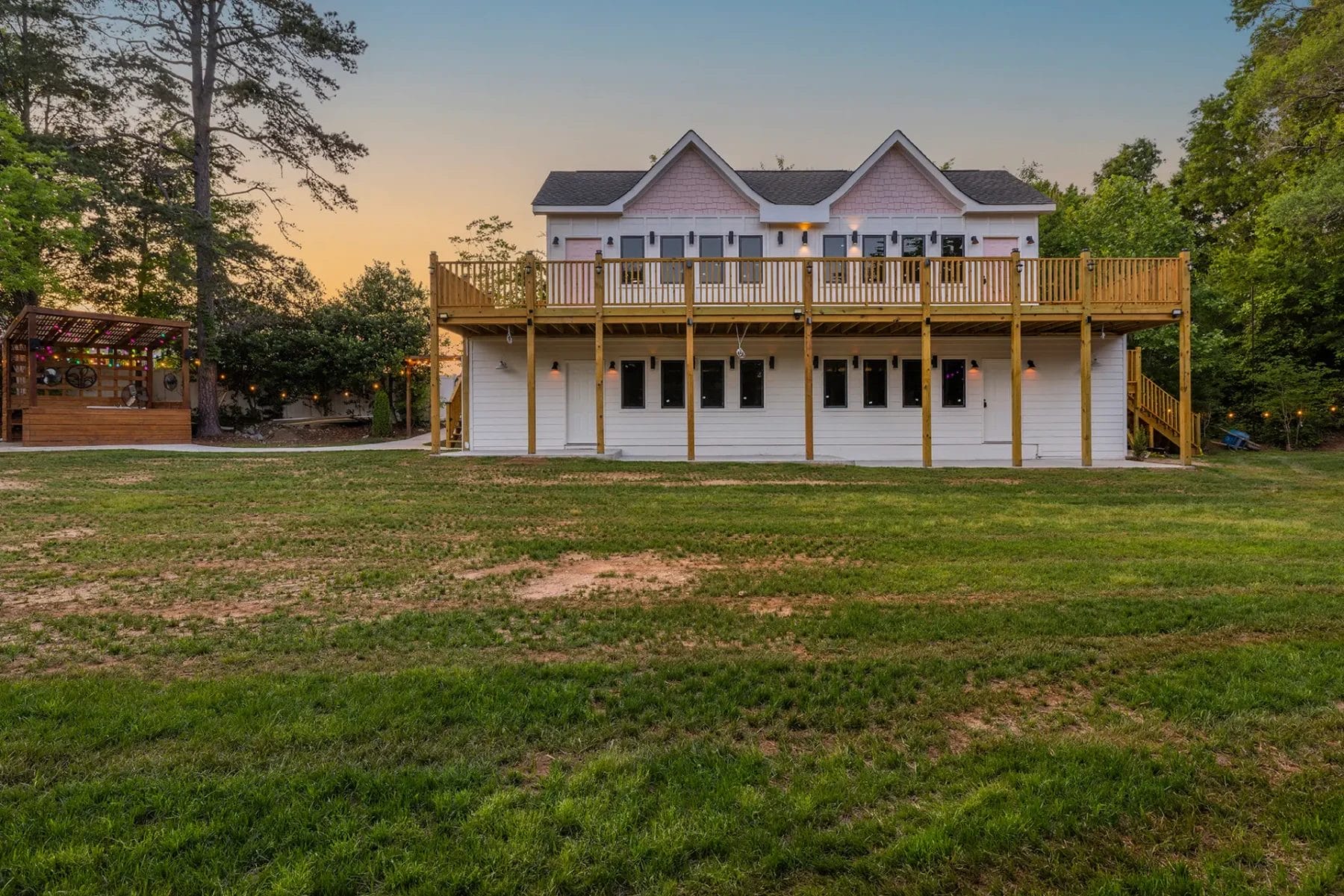 A two-story house with a large wooden deck and railing, set on a spacious grassy yard at sunset, surrounded by trees under a clear sky. Huntersville event venue