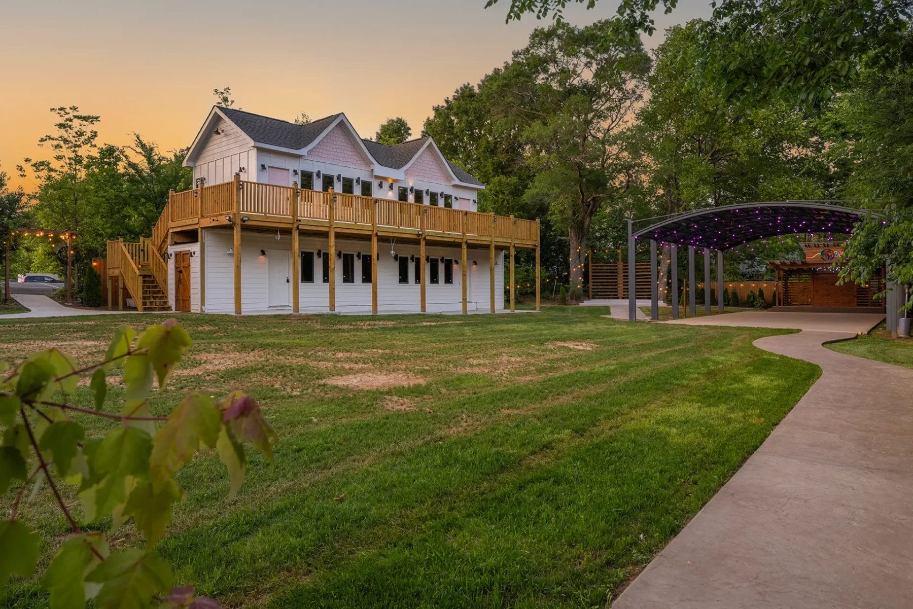 A large white house with a wooden balcony stands next to a green lawn. A curved sidewalk leads to a covered outdoor stage with purple lights. Trees surround the area at sunset. Huntersville event venue