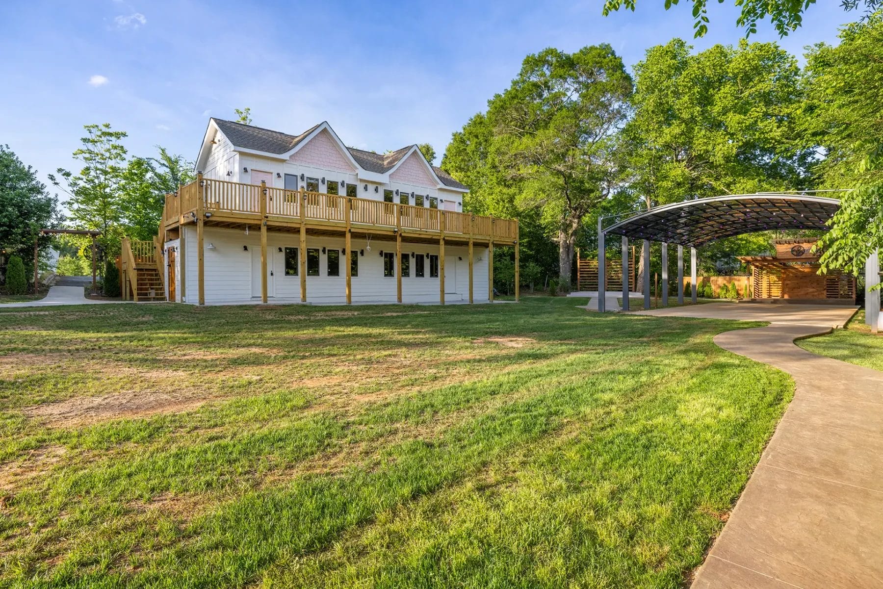 A two-story white house with a large wooden deck and balcony sits on a grassy yard, surrounded by trees. A curved paved walkway leads to a covered carport on the right side of the image. Huntersville event venue