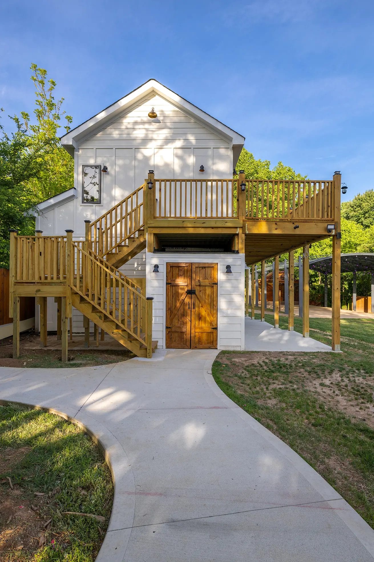 A two-story white building with a wooden door, wraparound wooden deck, and exterior stairs, surrounded by green trees and a curved concrete pathway leading to the entrance. Huntersville event venue