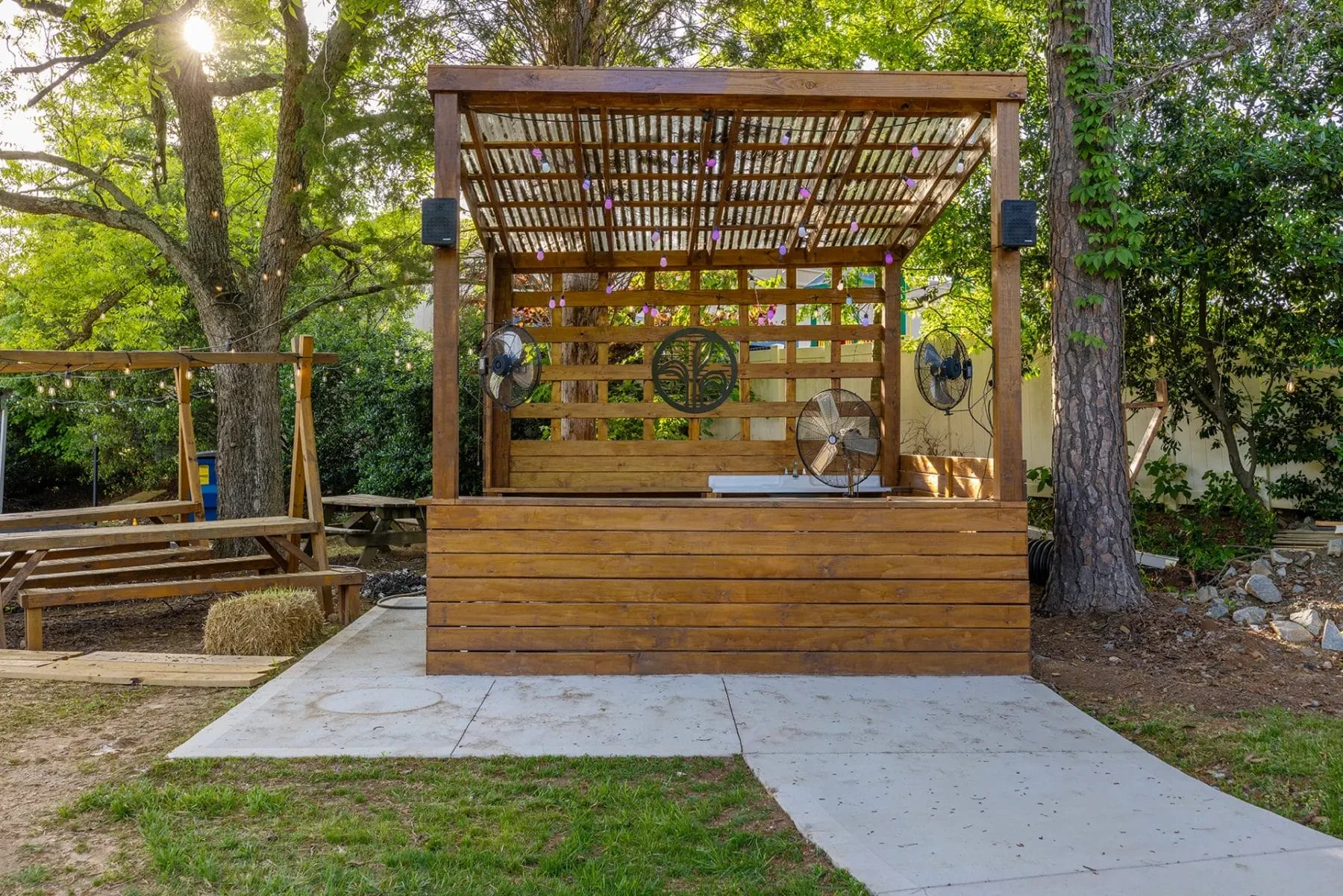 A small outdoor wooden stage with a slatted roof and side fans, surrounded by trees and greenery, with a concrete path leading up to it and benches nearby. Sunlight filters through the branches. Huntersville event venue