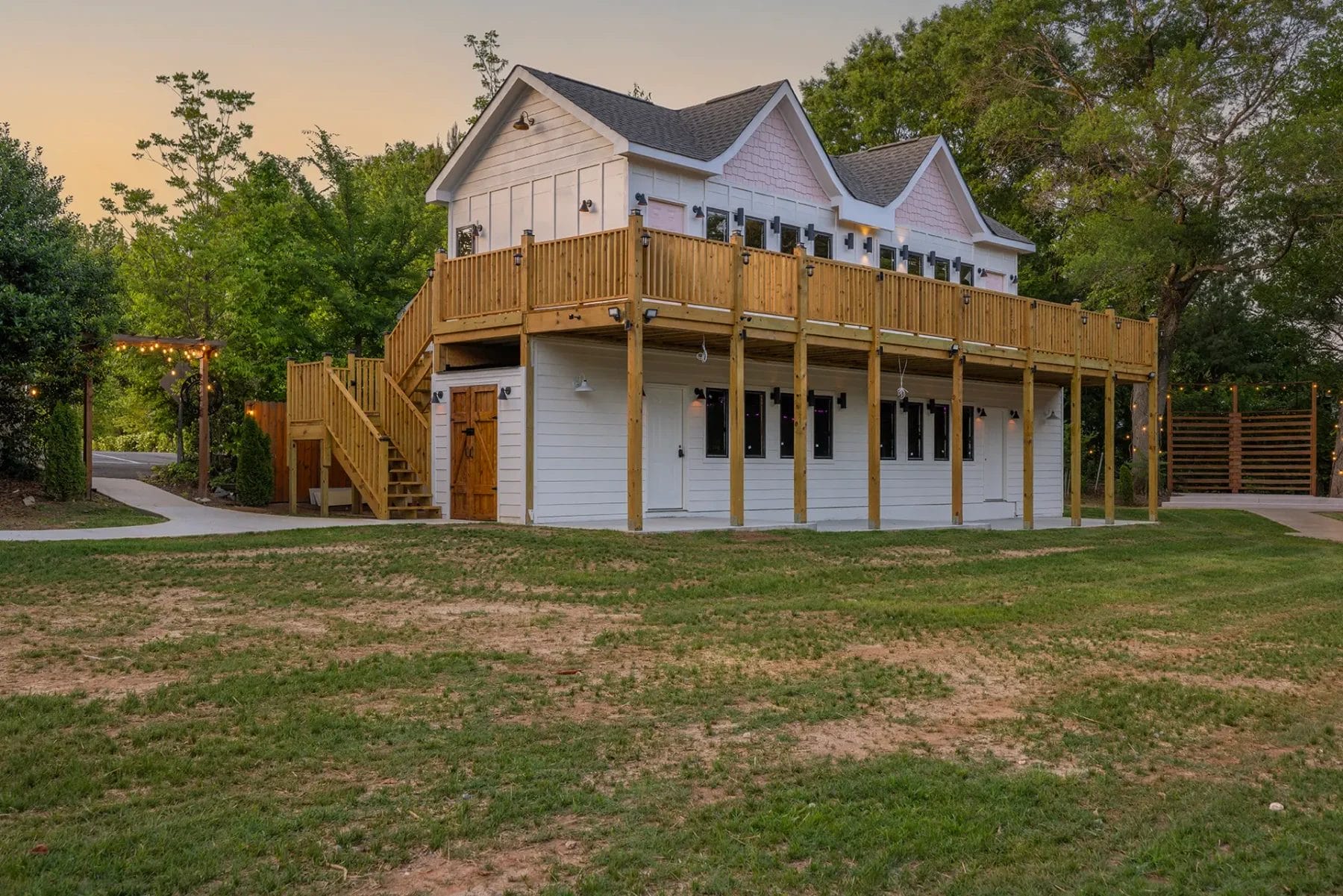 First Look: Exciting New Transformations at Manjal Manor 3 Two-story white house with a large wooden deck and staircase, surrounded by a grassy lawn and trees. The upper floor features windows and railing, while the ground floor has multiple doors and windows. Huntersville event venue