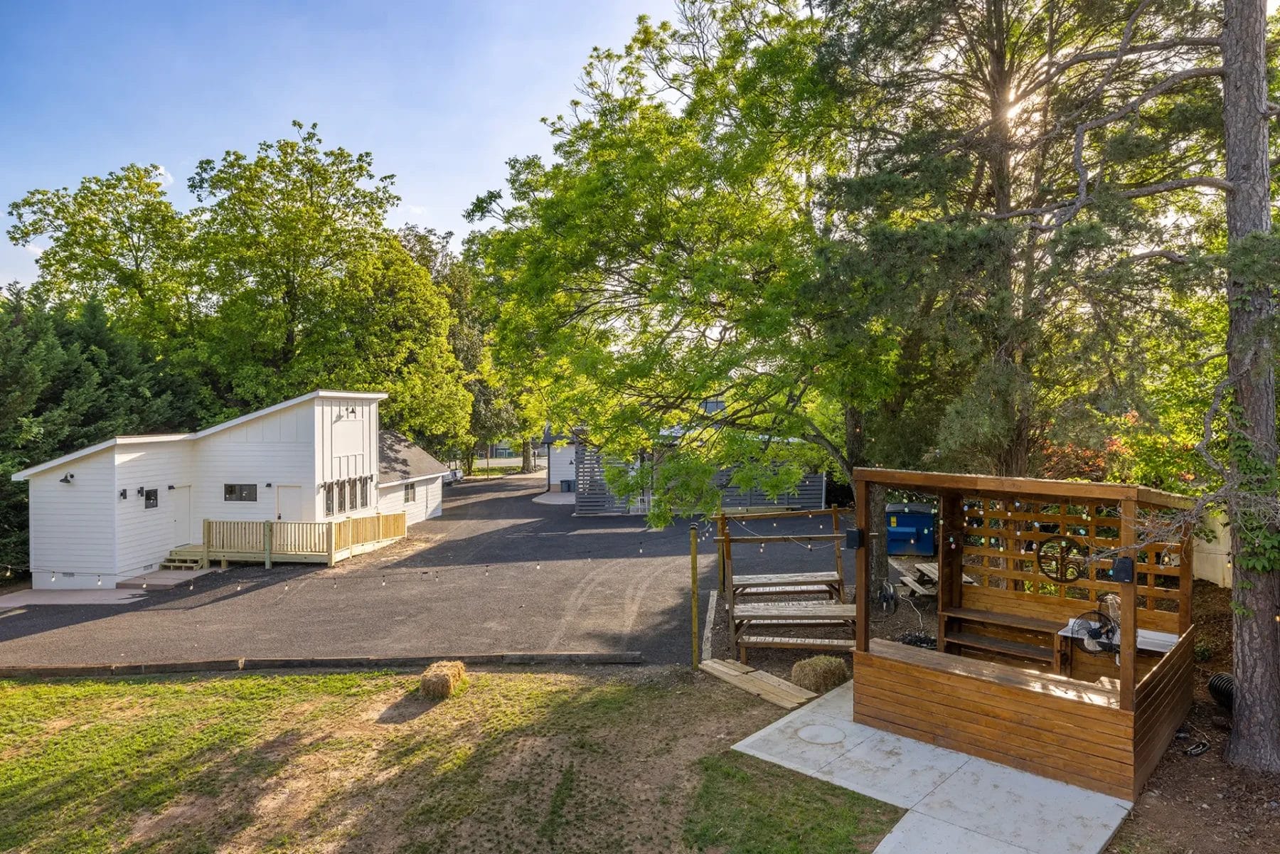 First Look: Exciting New Transformations at Manjal Manor 42 A sunny outdoor scene with a white building, paved driveway, trees, and a wooden deck with seating and a bicycle. Shadows from the trees stretch across the ground in a peaceful, open area. Huntersville event venue