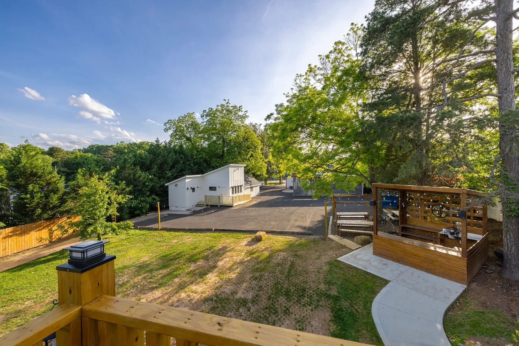 View from a wooden deck overlooking a paved parking lot, a white building, trees, and a small fenced seating area with benches, on a sunny day. Huntersville event venue