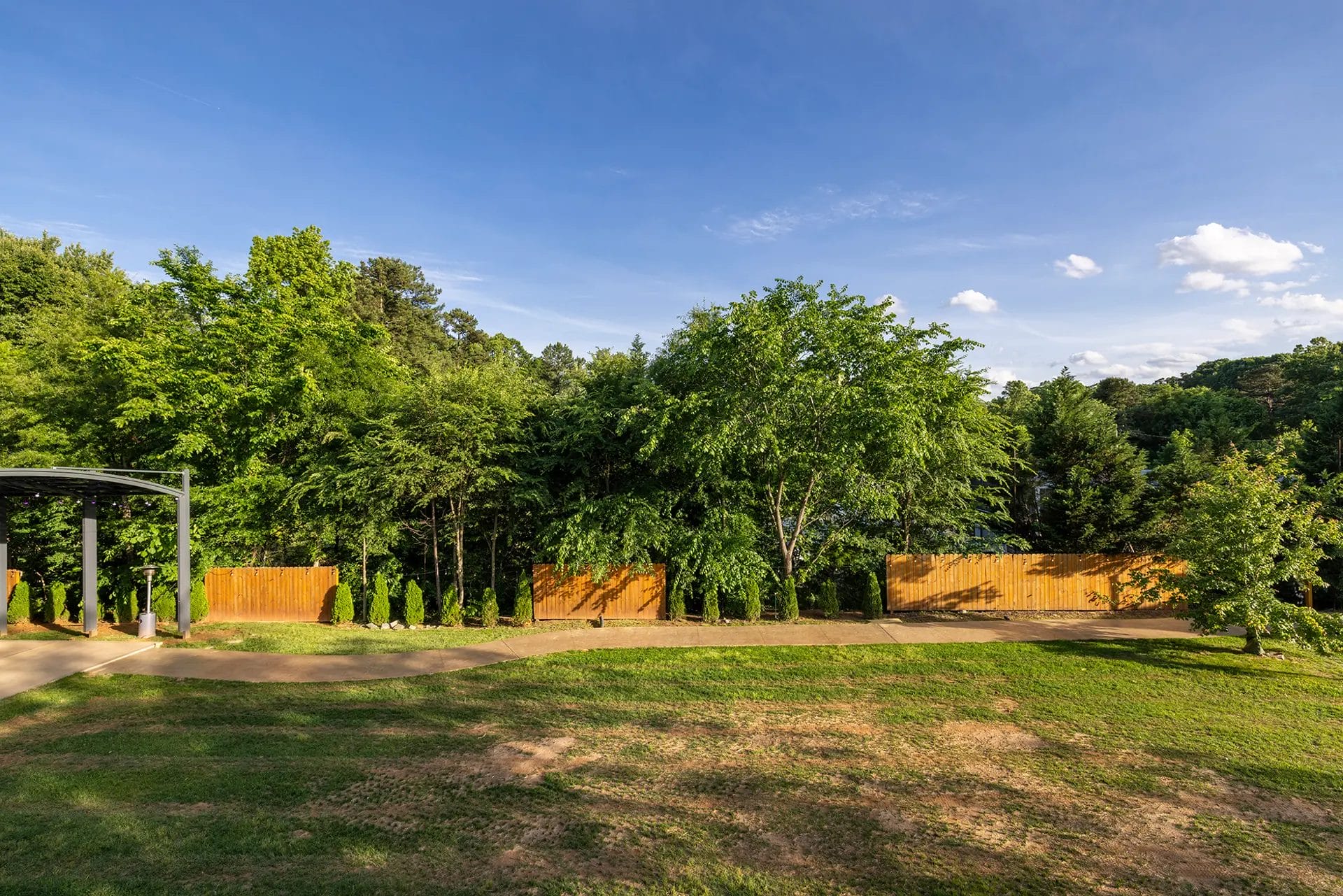 A backyard with green grass, a paved walkway, tall leafy trees, and a wooden fence under a blue sky with scattered clouds. Huntersville event venue