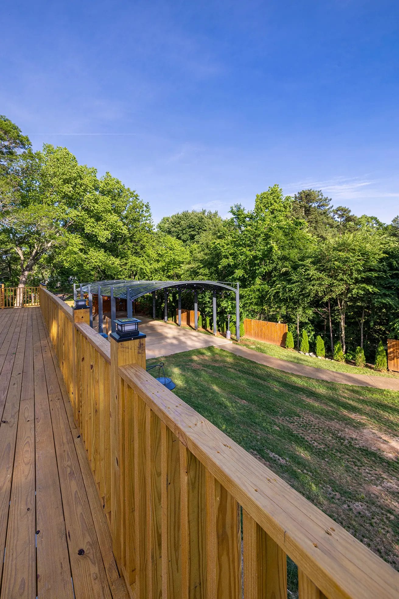 First Look: Exciting New Transformations at Manjal Manor 25 A wooden deck with a railing overlooks a grassy yard and a covered parking area, surrounded by tall green trees under a clear blue sky. Huntersville event venue