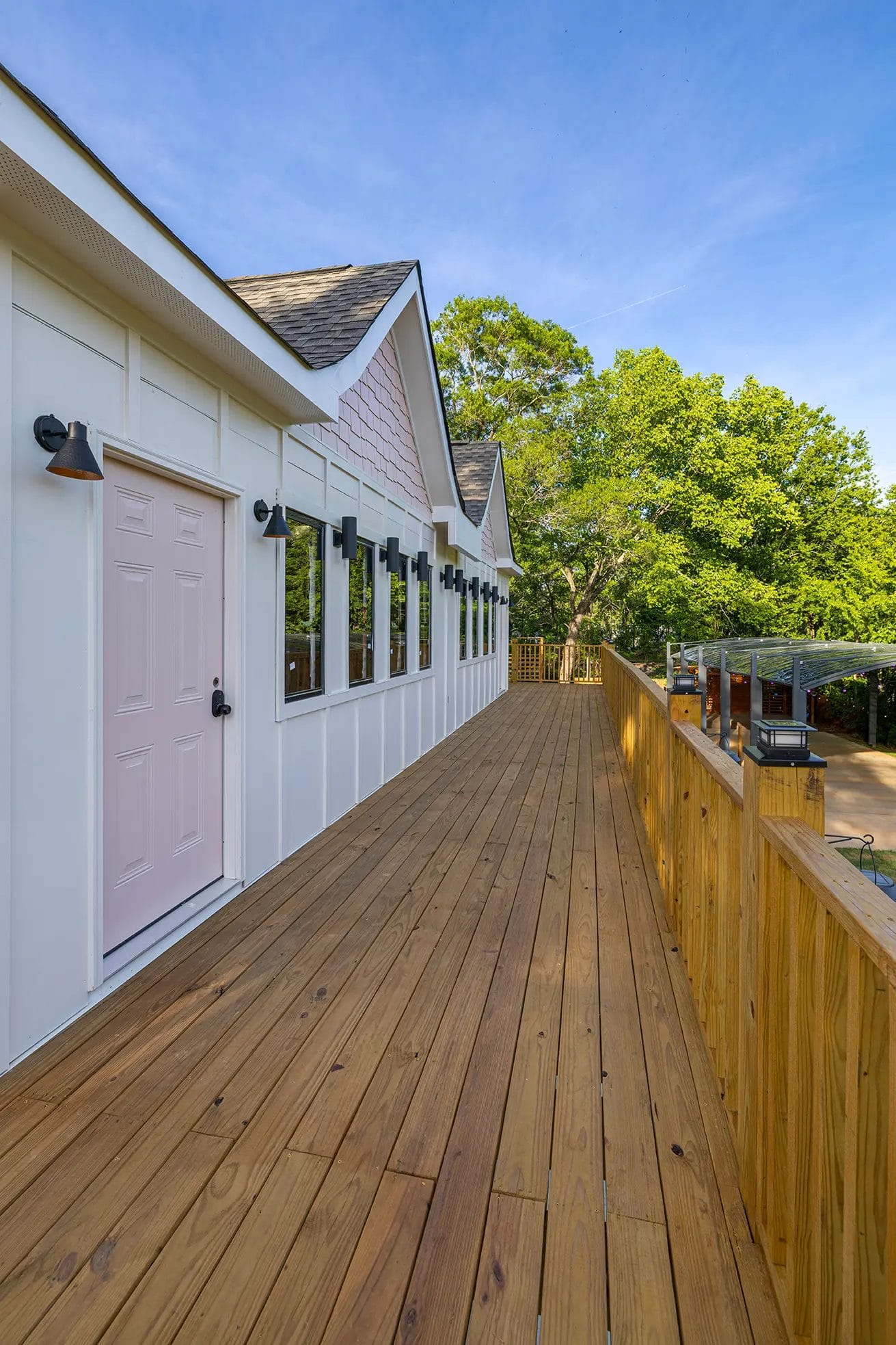 A long wooden deck with a railing runs alongside a white house with black-trimmed windows and a light pink door, surrounded by green trees under a clear blue sky. Huntersville event venue