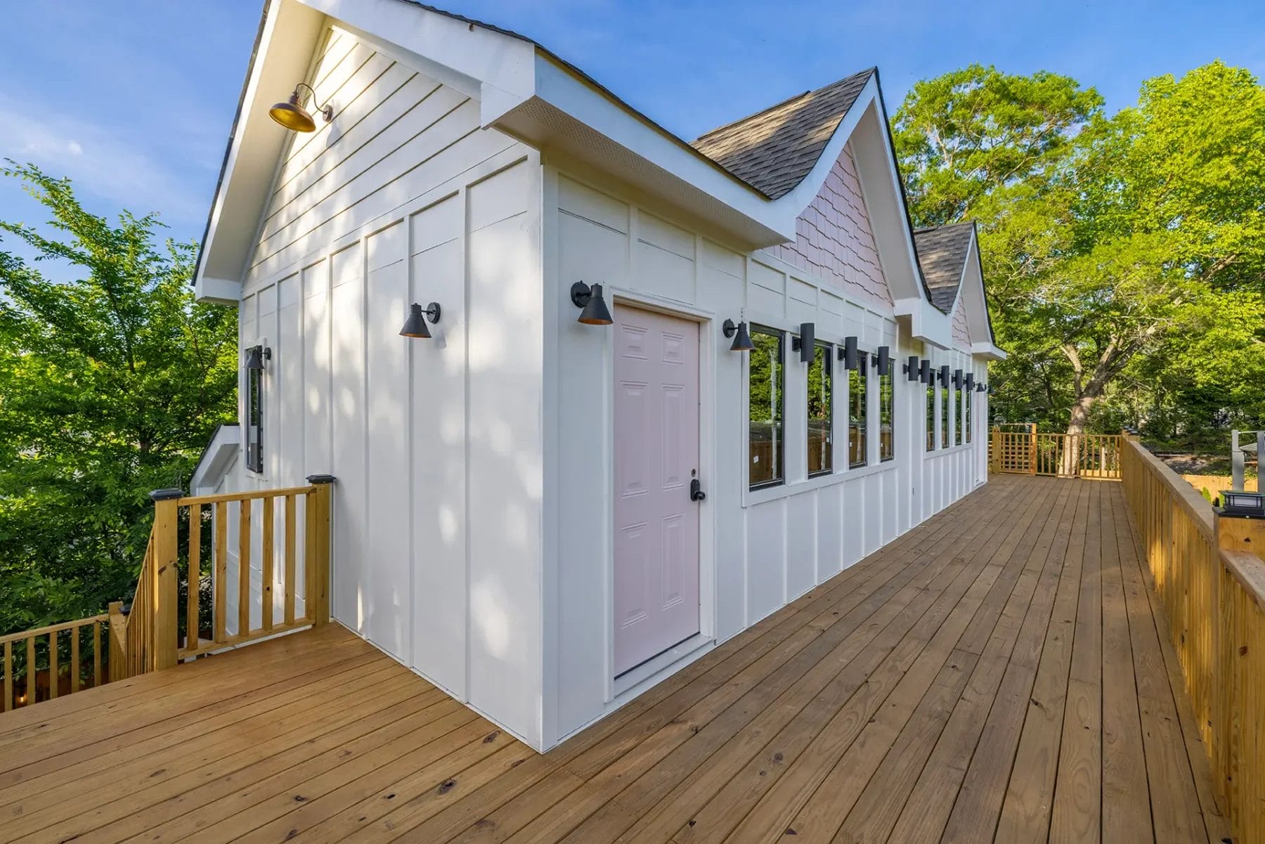 A modern white building with large windows and a light pink door sits on a spacious wooden deck surrounded by green trees under a blue sky. Huntersville event venue