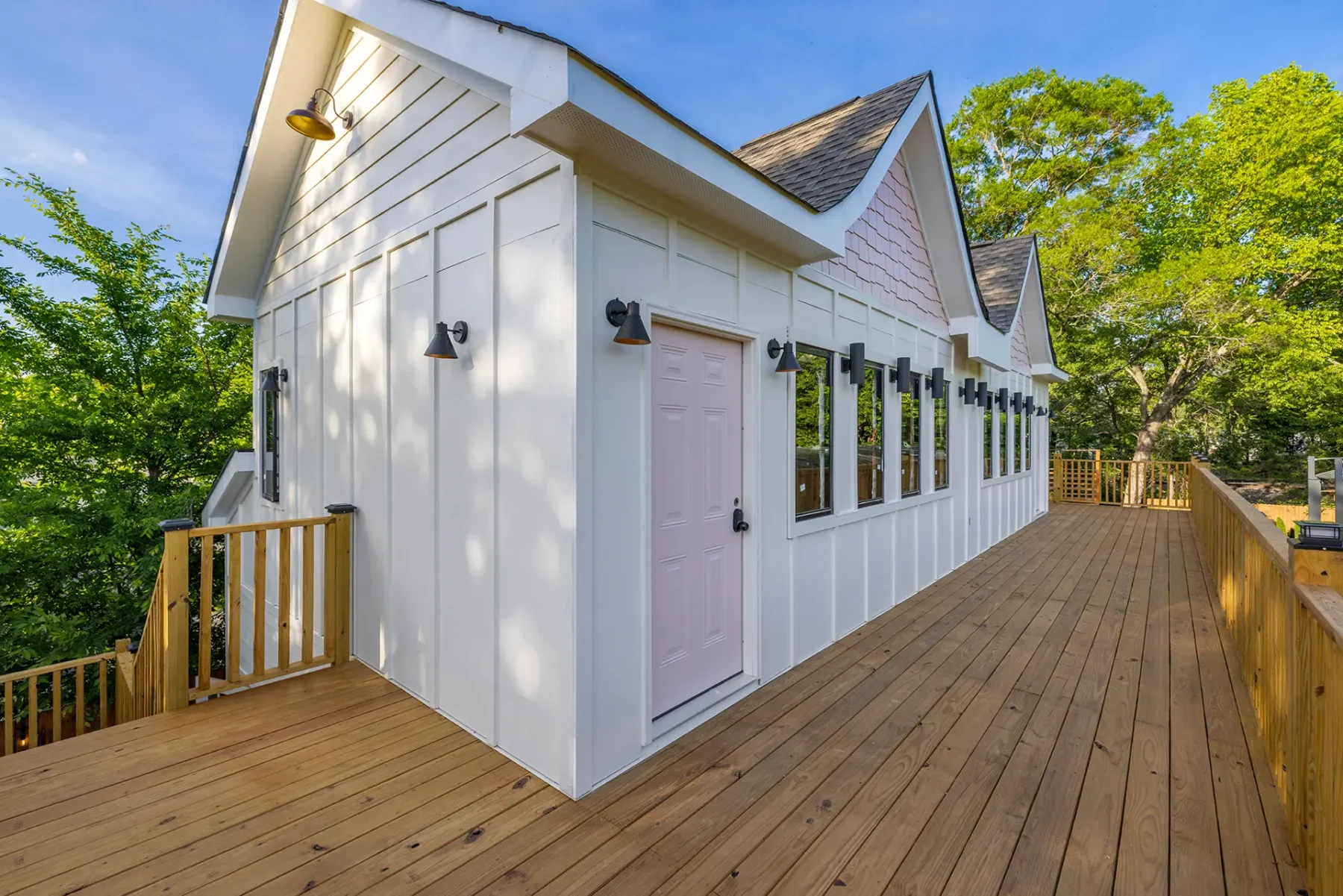 First Look: Exciting New Transformations at Manjal Manor 24 A modern white building with large windows and a light pink door sits on a spacious wooden deck surrounded by green trees under a blue sky. Huntersville event venue
