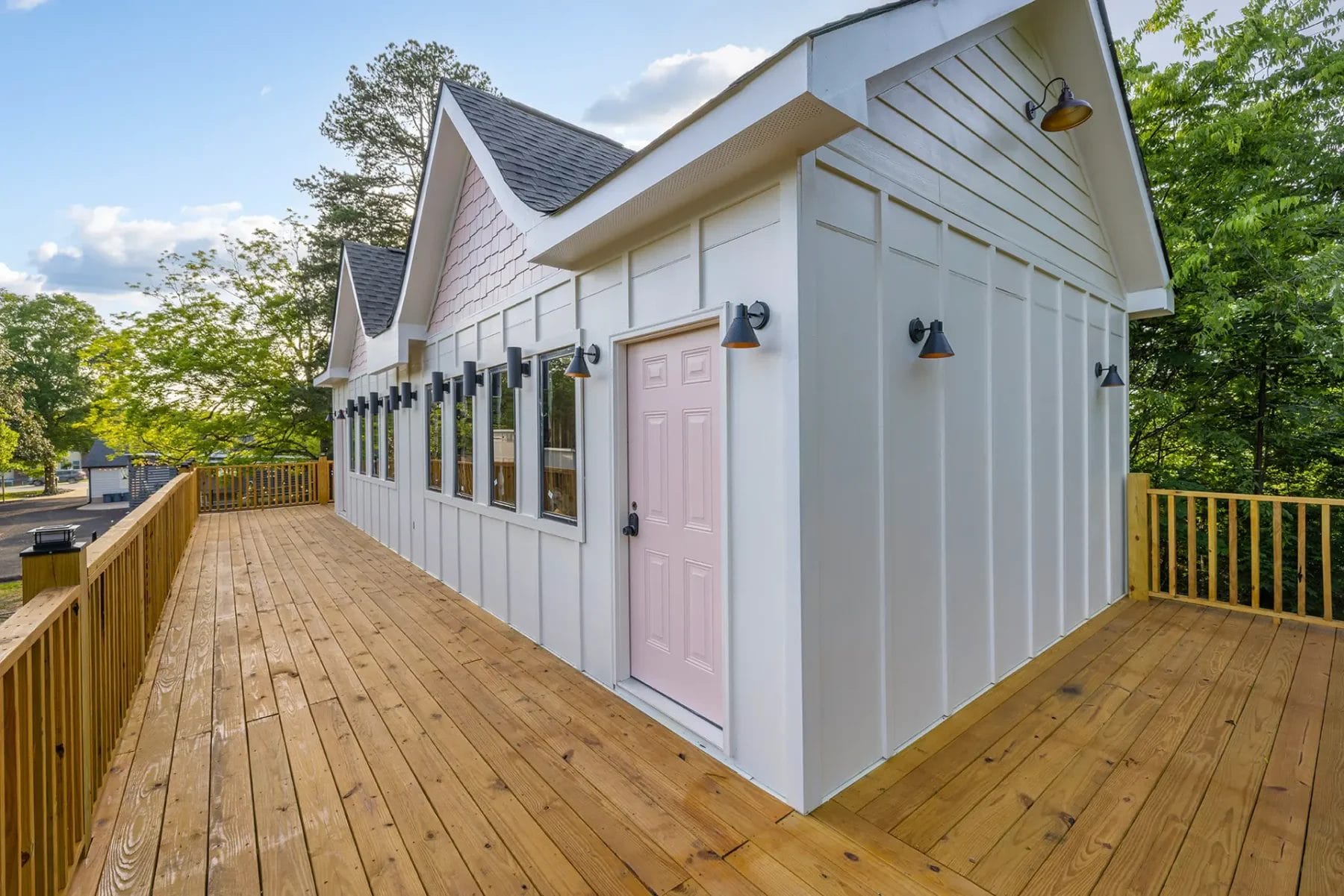 A modern white house with a light pink door, black outdoor sconces, large windows, and a spacious wooden deck surrounded by trees. Huntersville event venue