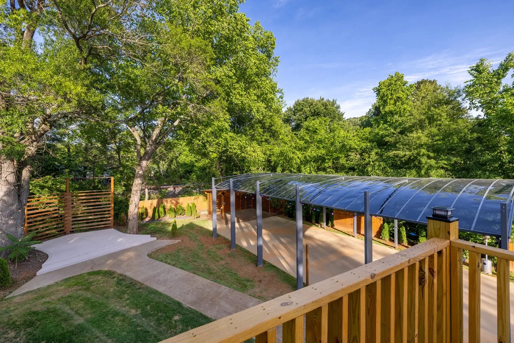 View of a backyard with a modern covered carport, paved driveway, wooden railing in the foreground, lush green trees, and a clear blue sky. Huntersville event venue