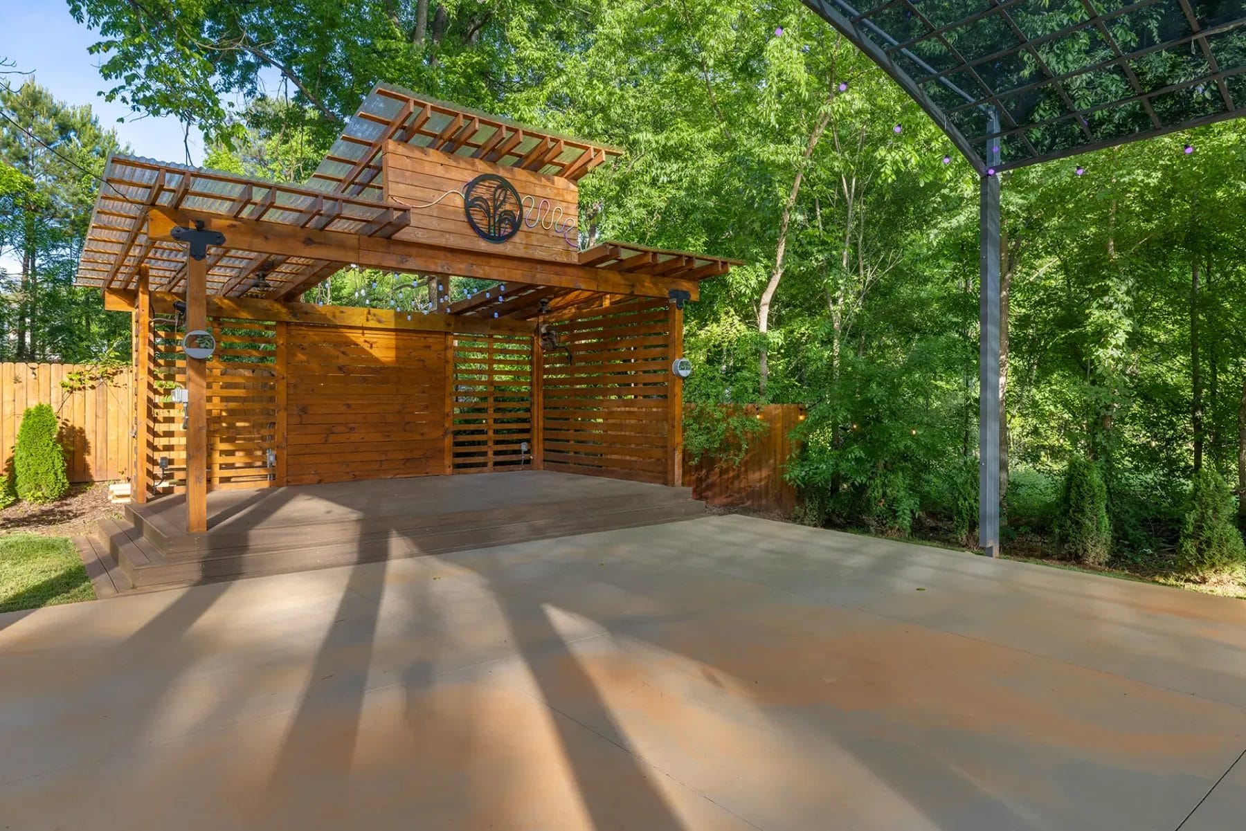 First Look: Exciting New Transformations at Manjal Manor 72 Outdoor wooden stage with a pergola roof, surrounded by trees and greenery. The stage is empty, with sunlight casting shadows on the adjacent open concrete area. A wooden fence is visible on the left. Huntersville event venue