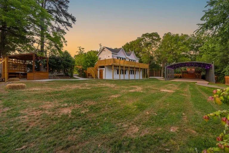 A large grassy yard with a white house featuring a wraparound porch, a wooden stage area to the right with string lights, and a wooden gazebo structure to the left, surrounded by tall trees at sunset. Huntersville event venue