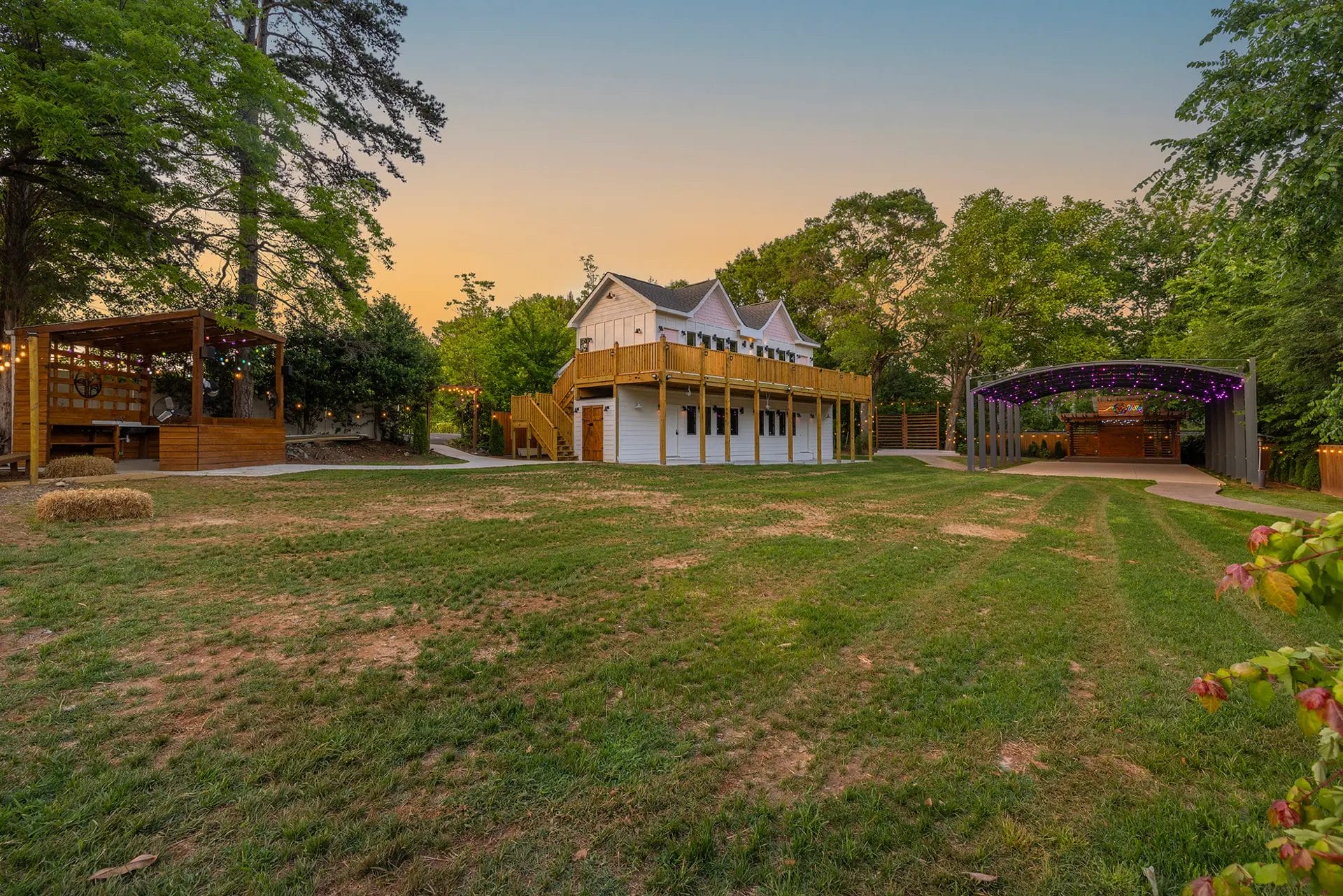 A large grassy yard with a white house featuring a wraparound porch, a wooden stage area to the right with string lights, and a wooden gazebo structure to the left, surrounded by tall trees at sunset. Huntersville event venue