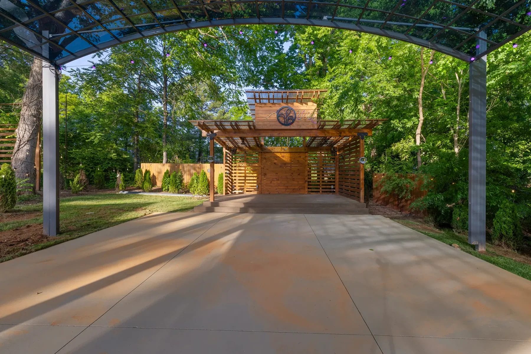A modern carport with a wooden structure and metal roof stands at the end of a wide concrete driveway, surrounded by green trees and a landscaped yard under a sunny sky. Huntersville event venue