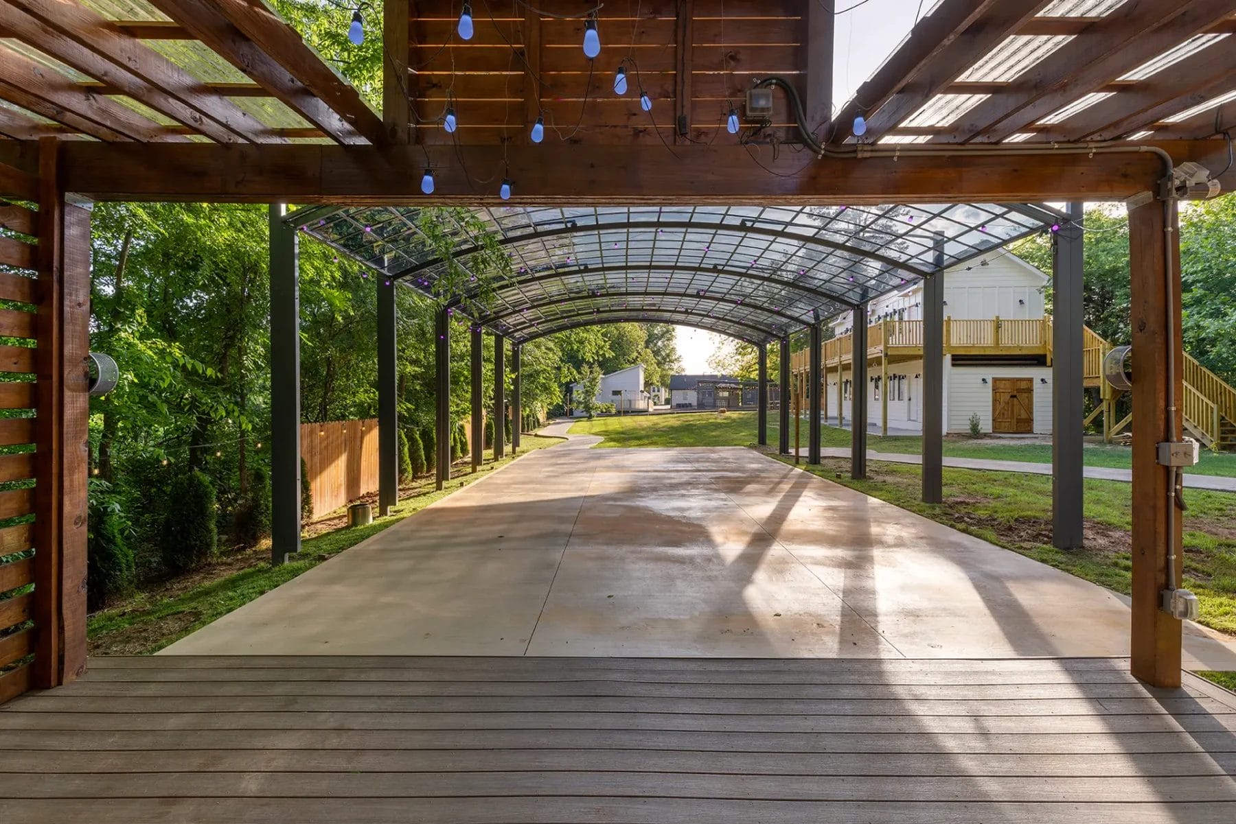 View from a wooden deck under a pergola with hanging blue lights, looking out onto a covered walkway with a clear roof, surrounded by greenery and a two-story building in the background. Huntersville event venue