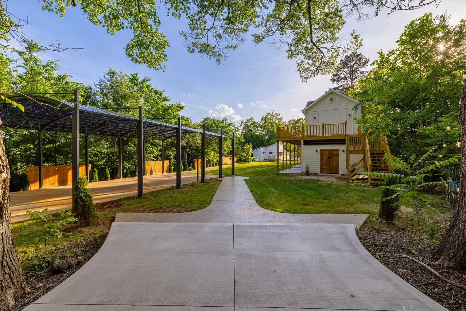 A paved path leads to a small white two-story building with a wooden deck, surrounded by trees and greenery. On the left, there is a covered structure with a metal roof and support poles. Huntersville event venue