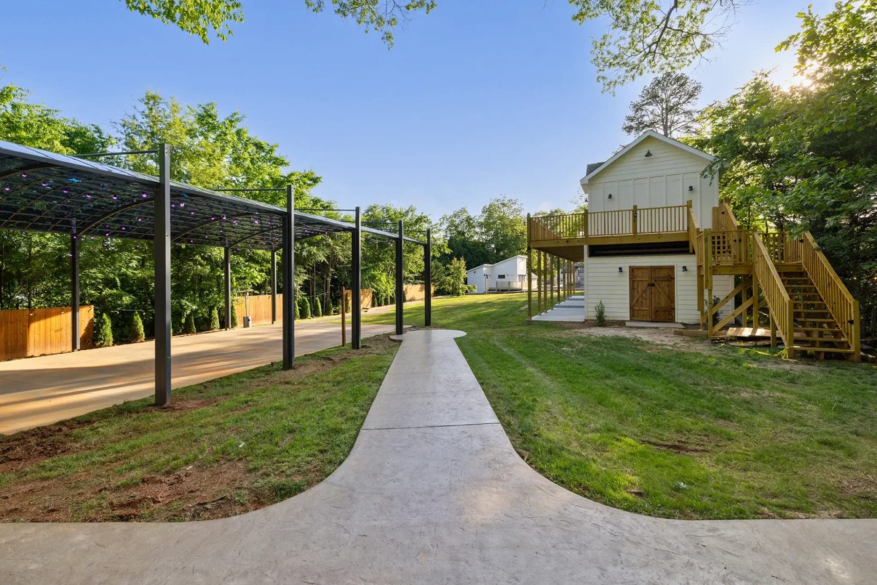 A paved walkway leads to a wooden two-story building with exterior stairs and balcony, set in a grassy yard. On the left, there is a covered open structure with a metal roof and string lights. Trees and fenced area surround the space. Huntersville event venue