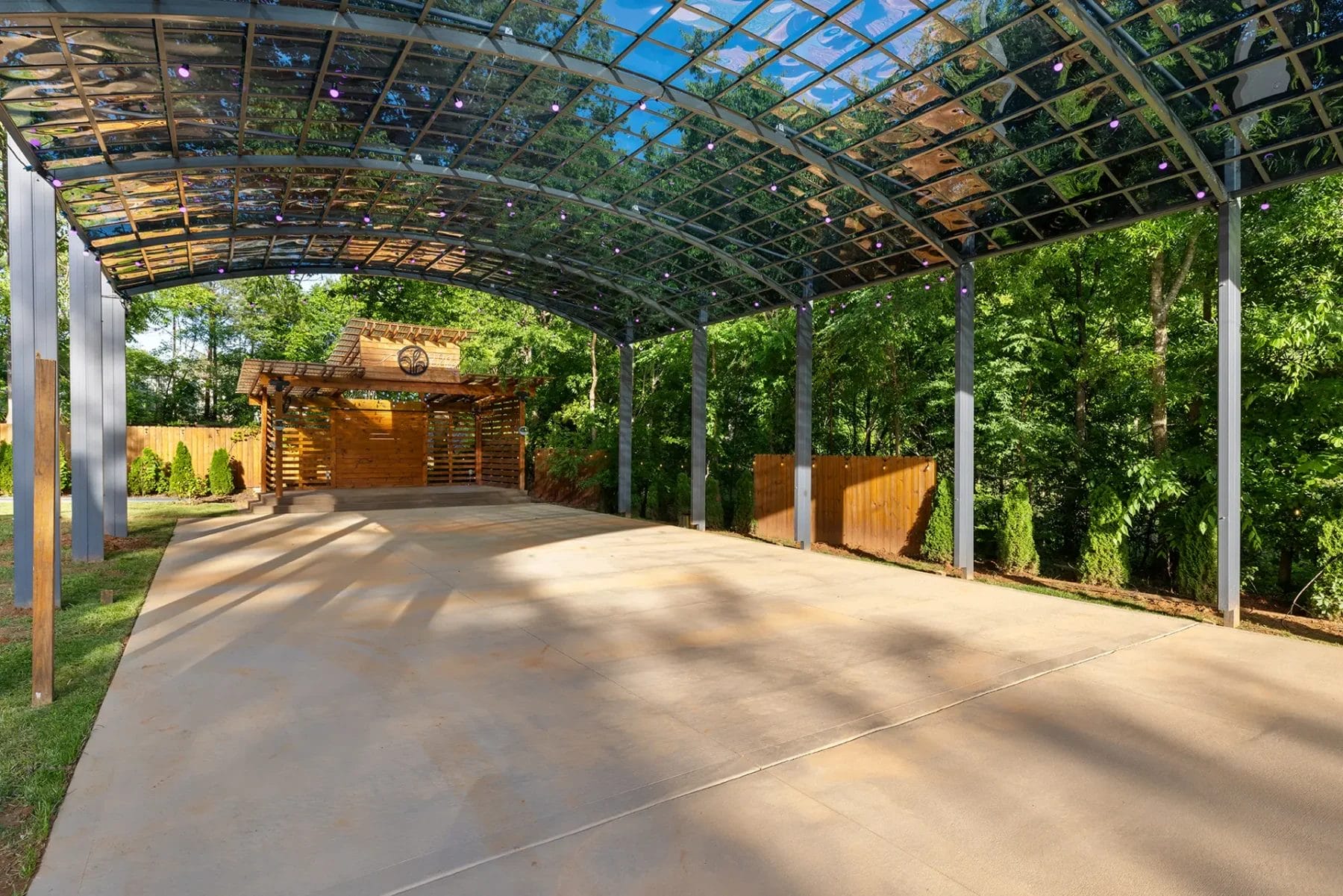 A covered outdoor pavilion with a curved, transparent roof and open sides, set on a large concrete pad, surrounded by trees and greenery, with a wooden structure at the far end. Huntersville event venue