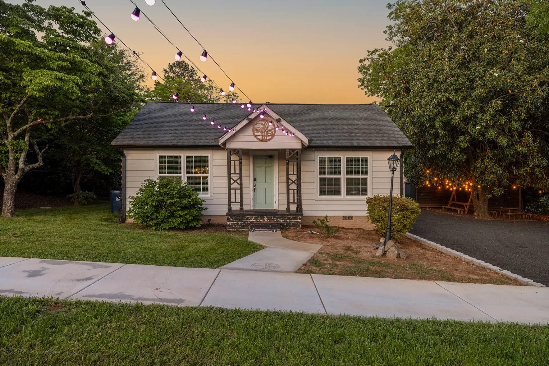 A small, light-colored house with string lights overhead, surrounded by trees and greenery at sunset. A paved walkway leads to the front door, and outdoor lights create a cozy atmosphere. Huntersville event venue