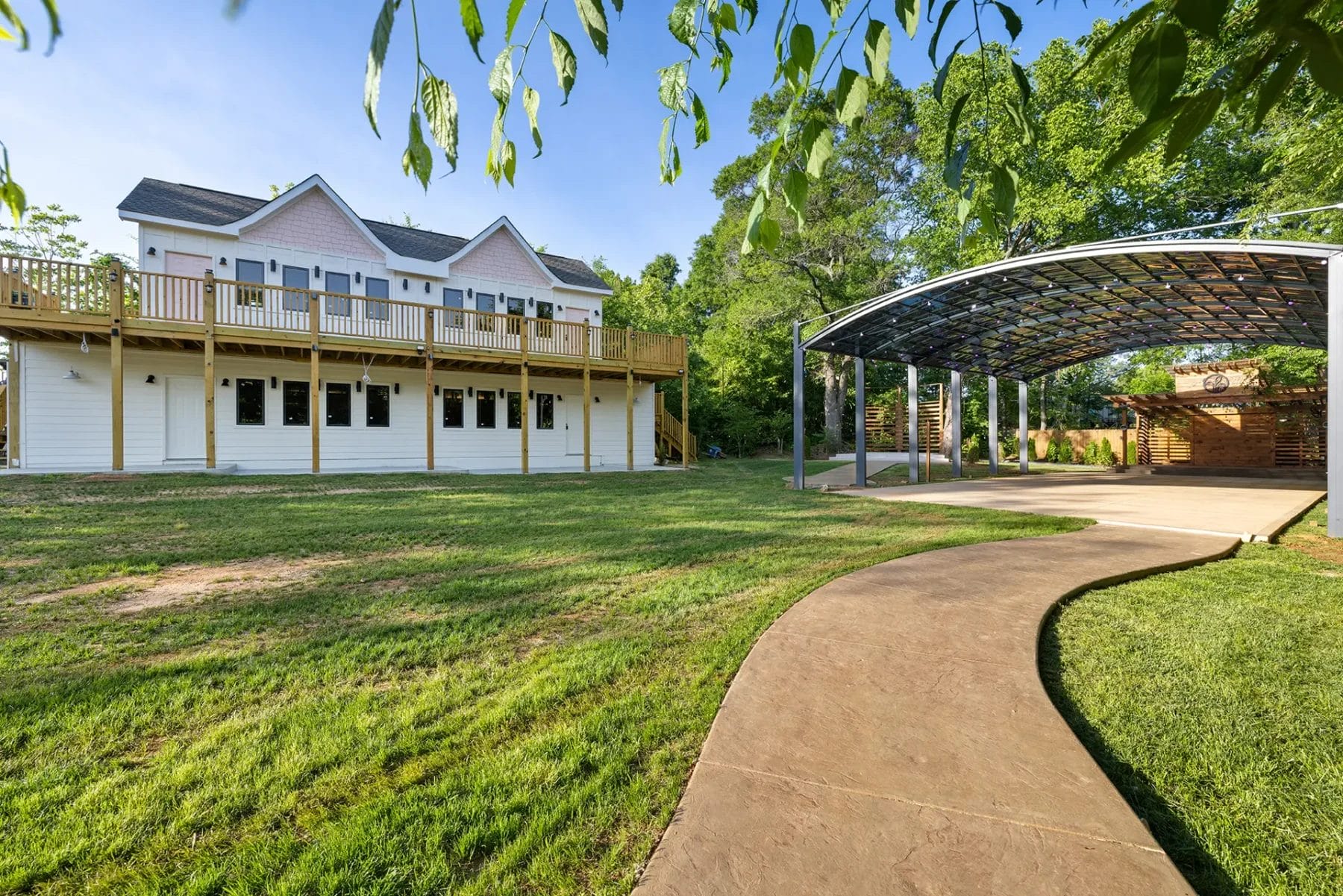 A white two-story house with a large wooden deck stands next to a covered outdoor pavilion, surrounded by green grass, trees, and a curved concrete walkway under sunny skies. Huntersville event venue