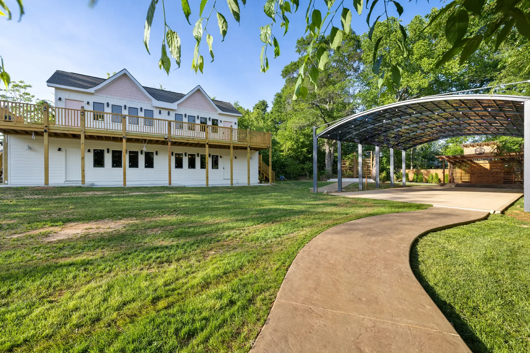 First Look: Exciting New Transformations at Manjal Manor 10 A white two-story house with a large wooden deck stands next to a covered outdoor pavilion, surrounded by green grass, trees, and a curved concrete walkway under sunny skies. Huntersville event venue