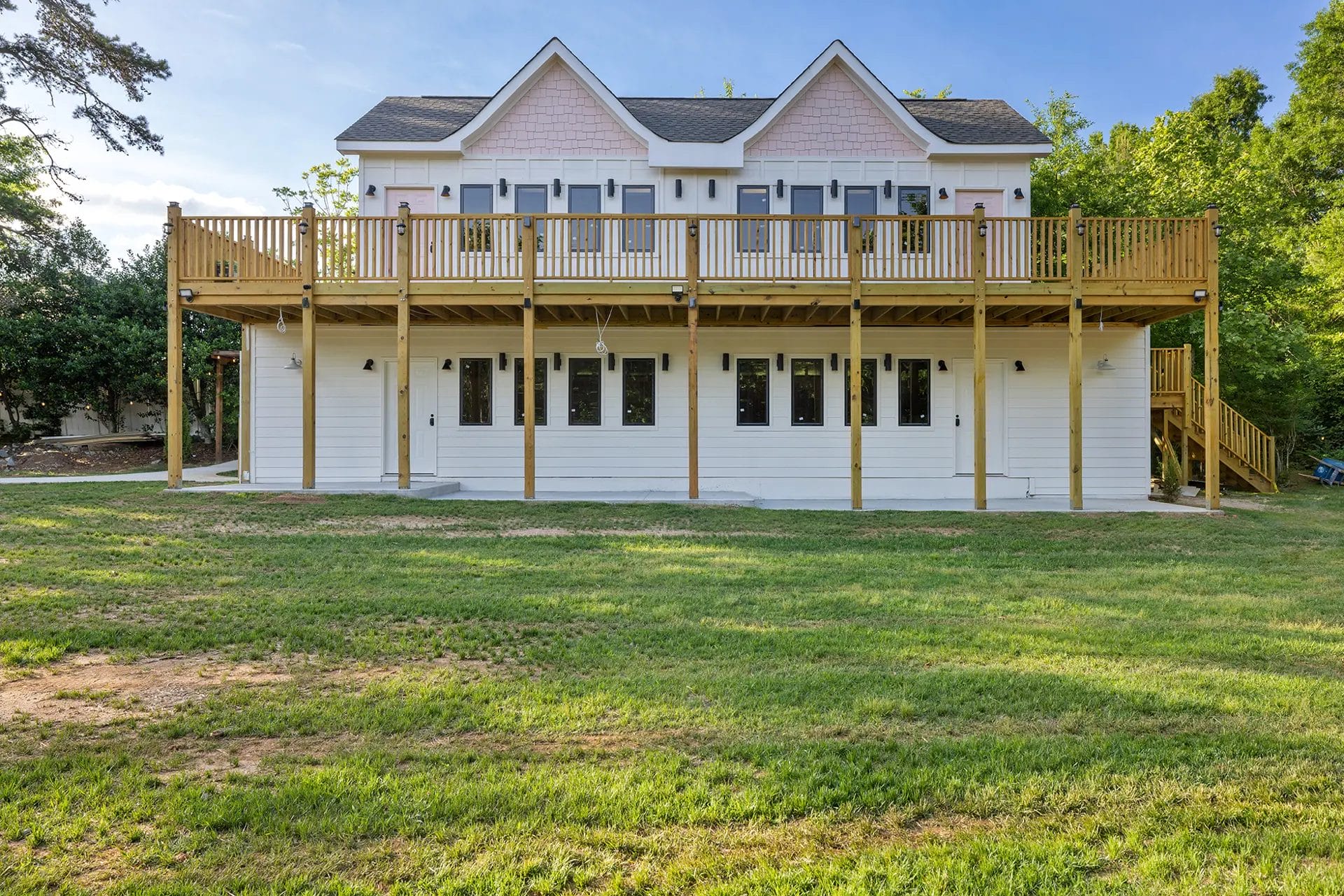Two-story white house with pink accents, large upper wooden balcony with railings, multiple glass doors and windows, and surrounded by a grassy yard and trees. Huntersville event venue