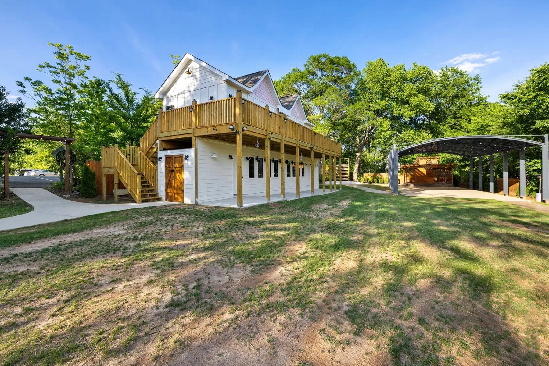 A two-story white house with a large wooden deck and staircase, surrounded by green trees. There's a covered parking area on the right and a patchy grassy yard under a clear blue sky. Huntersville event venue