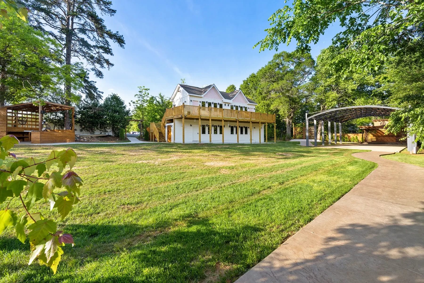 A large two-story white house with a wraparound deck is surrounded by green lawns, trees, and wooden gazebos. A curved concrete path leads through the landscaped yard under a blue sky. Huntersville event venue
