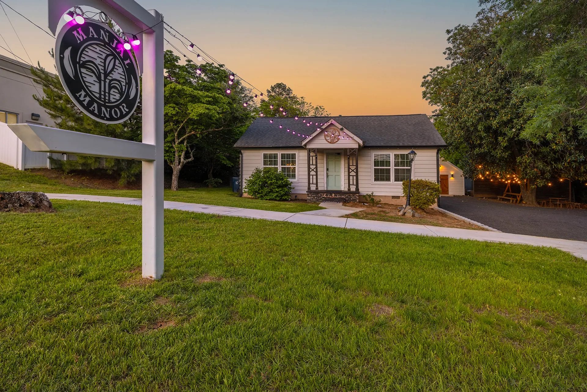 A small white house with a porch sits among green trees at sunset. Pink string lights connect a white "Manor" sign in the yard to the house. The sky is orange and purple, casting a warm glow over the scene. Huntersville event venue