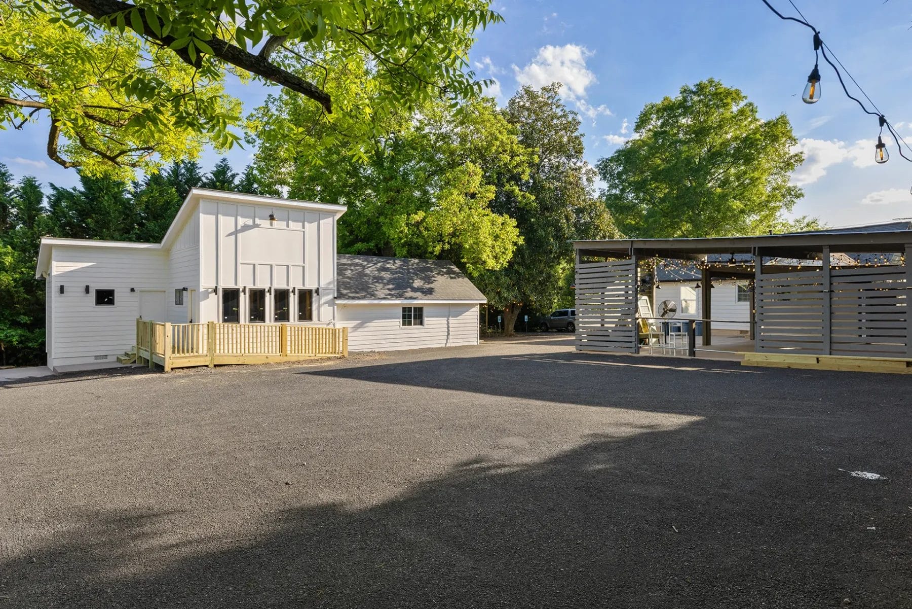 A paved outdoor area with a modern white building, a smaller structure, trees, and a covered patio with string lights and seating on a sunny day. Huntersville event venue