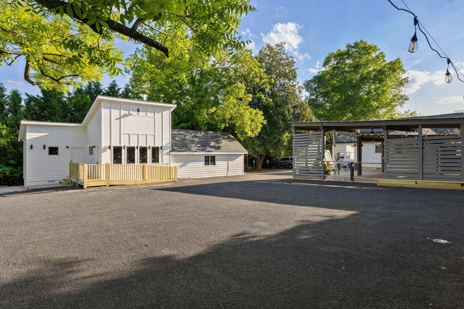 First Look: Exciting New Transformations at Manjal Manor 35 A paved outdoor area with a modern white building, a smaller structure, trees, and a covered patio with string lights and seating on a sunny day. Huntersville event venue