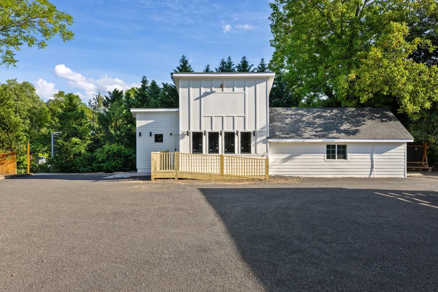 A modern white building with large windows, a wooden accessibility ramp, and an asphalt parking area, surrounded by trees under a bright blue sky. Huntersville event venue