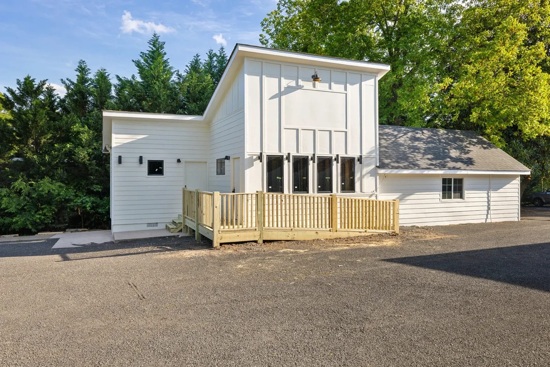 A modern white house with large windows, a sloped roof, and a wooden ramp leads to the entrance. The home is surrounded by trees and is adjacent to a paved driveway. Huntersville event venue